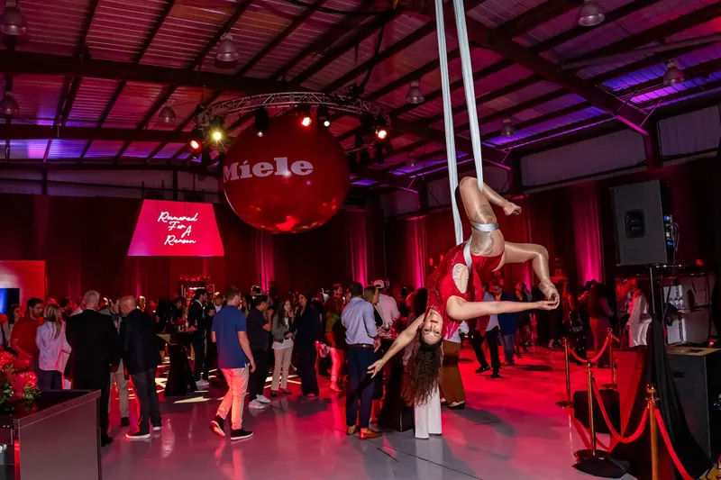 Aerialist performing on silks above guests at a red-lit Miele brand activation inside a draped industrial space